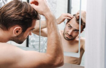Worried young shirtless man examining his hair while looking at the bathroom mirror